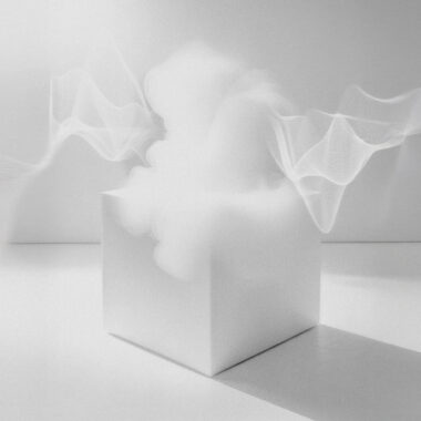 Fotografia czarno biała. Puste pomieszczenie z białymi ścianami i białą podłogą. Na środku stoi biały sześcian, z którego wydobywa się biały dym.