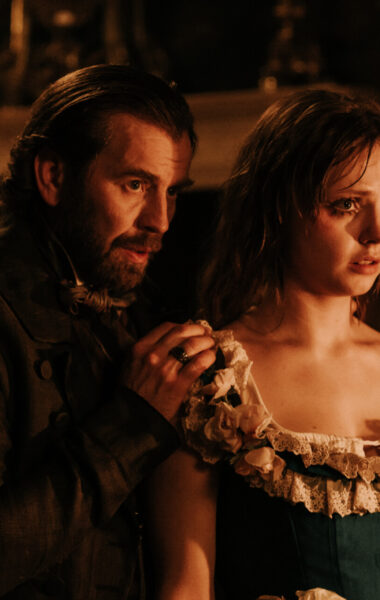 Kadr z filmu kostiumowego. dziewczyna i mężczyzna w starodawnych strojach stoją blisko siebie. Mężczyzna stoi nieco za dziewczyną, trzymając dłoń na jej ramieniu. Za nimi niewyraźne elementy architektury lub mebli wyłaniające się z ciemności.