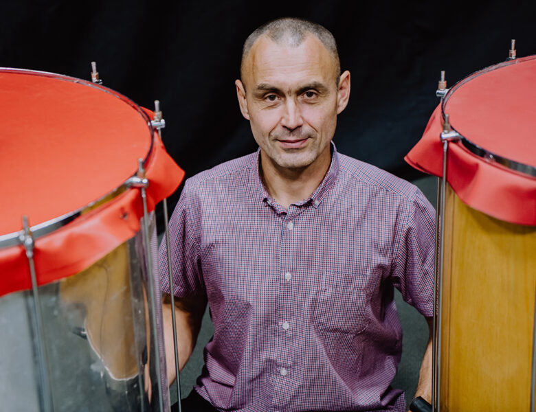 Między dwoma bębnami siedzi mężczyzna. Patrzy prosto w obiektyw. Za nim czarne tło.